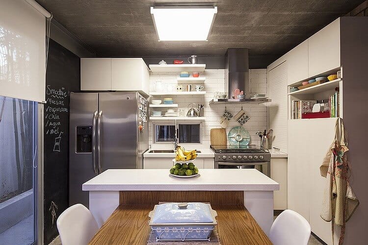 The small and homey industrial kitchen has a white kitchen island that has wooden table attached to it serving as a dining table. The white peninsula is dominated by the large fridge that matches with the gray concrete ceiling.