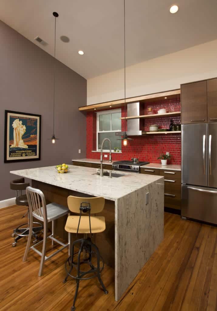 The simplicity of this kitchen is elevated to a higher level with its combination of different hues. It has a white shed ceiling, gray wall and hardwood flooring. The white marble waterfall kitchen island stands out against this as well as the red backsplash tiles.