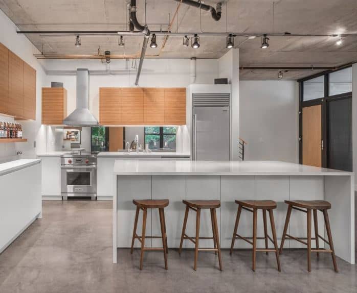 The gray concrete flooring of this industrial-style kitchen matches well with the gray concrete ceiling with exposed pipes and tubes supporting modern spot lights. This serves as a nice framing for the white modern kitchen island and peninsula.