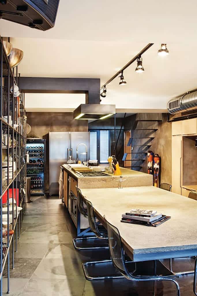 The kitchen island is made of concrete gray materials that extend to an attached dining area. This matches with the concrete flooring and walls that are dominated by a large shelving for storage complemented by the exposed vents of the ceiling.