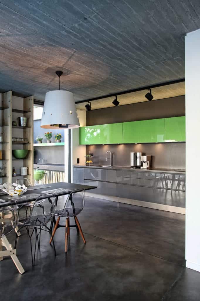 The concrete gray industrial-style flooring is a reflection of the ceiling that has a row of black spot lights mounted on it. This is matched with the dark gray walls and cabinets of the peninsula that makes the green floating cabinets stand out.