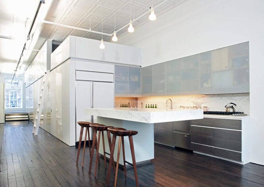 The simple and modern elements of the kitchen are given an industrial-style background with a distressed hardwood flooring, a high white ceiling with exposed pipes and pendant lights hanging over the thick white marble countertop of the kitchen island.