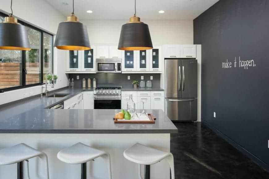 This industrial-style kitchen has a U-shaped peninsula with a gray countertop that complements the white kitchen cabinets and drawers and matches the stainless steel appliances. This is contrasted by a black wall with word artwork on it.