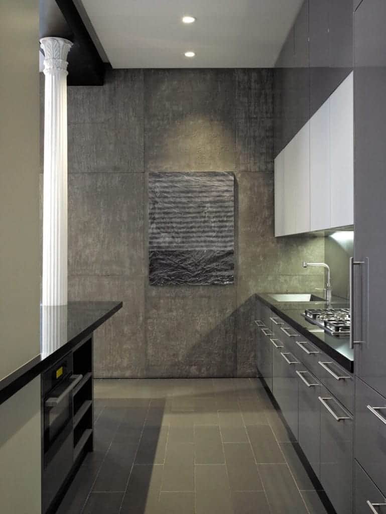 This small and narrow kitchen has a concrete gray flooring and wall that matches with the sleek gray cabinets and drawers of the kitchen island and peninsula with black countertops contrasted by the white pillar and ceiling.