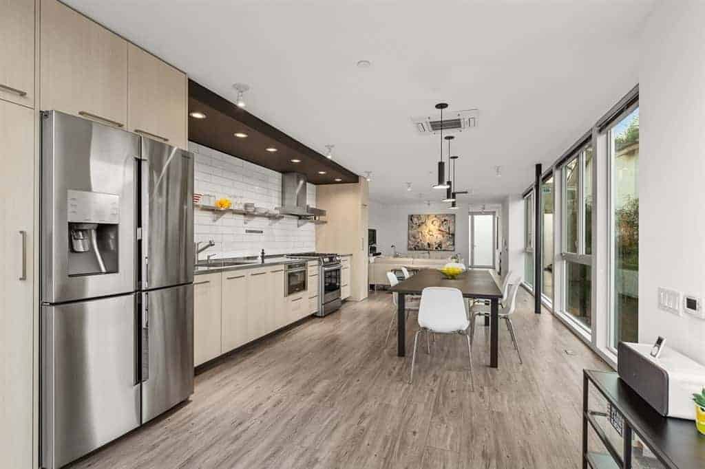 The kitchen peninsula's light gray countertop matches with the stainless steel appliances and complements the white backsplash arranged in a brick wall pattern that is contrasted by the dark wooden panel on the ceiling with recessed lights over the cooking area.