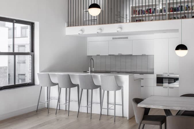 This industrial-style kitchen is right under the railings of the second level loft. The railings go well with the contrasting white walls that blend with the cabinets and drawers of the peninsula and kitchen island that is complemented by the gray countertop and backsplash.