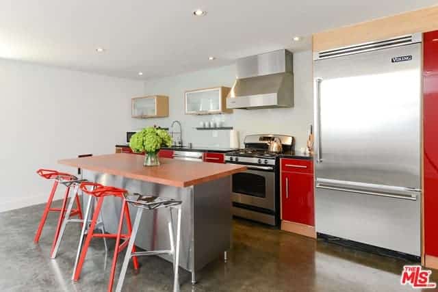 The sleek red elements of this industrial-style kitchen stands out against the stainless steel appliances and kitchen island that matches with the gray concrete flooring brightened by the white walls and ceiling.