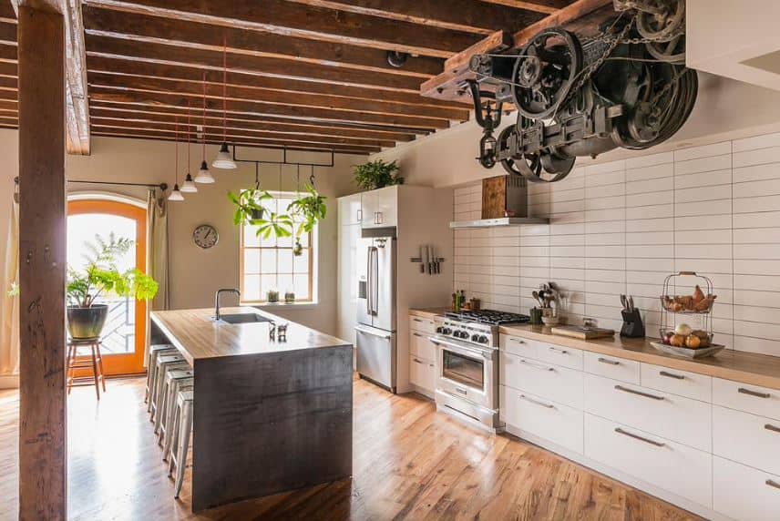 This kitchen has a hardwood flooring that matches with the wooden column supporting the wooden ceiling with exposed wooden beams. This ceiling is adorned with an old engine with gears and cogs that bring this kitchen a unique look.