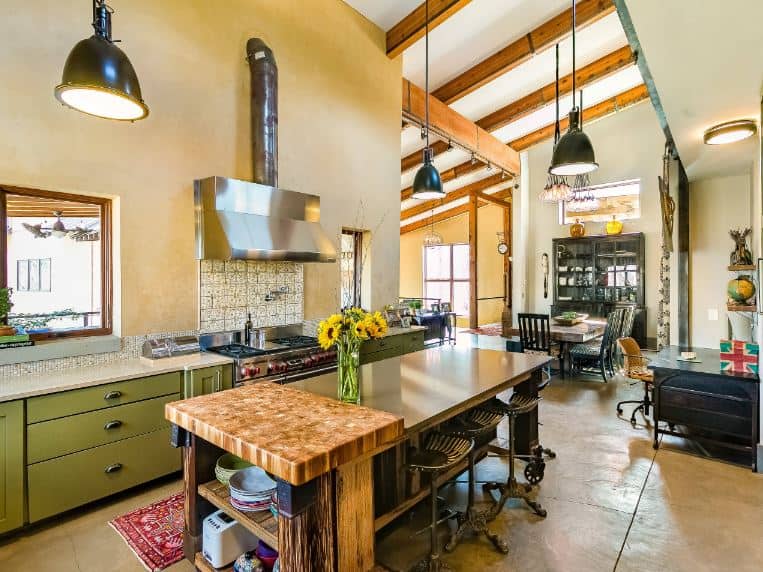 The textured beige flooring of this charming and homey industrial-style kitchen matches with the walls of the peninsula that has green wooden drawers and cabinets flanking the stainless steel stove-top oven topped with a matching vent embedded into the wall.