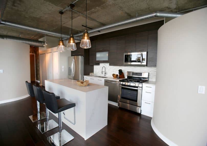 The dark hardwood flooring contrasts the white walls and the white kitchen island that matches the white peninsula. Above this peninsula are dark cabinets that go well with the concrete gray ceiling with exposed vents and ducts.