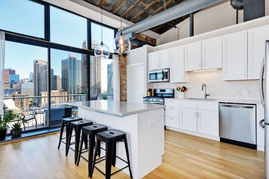 This bright kitchen has a high arched ceiling augmented by a large glass wall that offers a nice urban jungle scenery that brings in natural lights to the white kitchen island and peninsula that houses the stainless steel appliances.