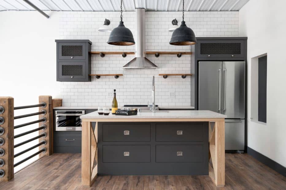 This is a second floor kitchen right beside the railings that have low wooden posts supporting metal rods. This pairs well with the dark gray kitchen cabinets of the island and peninsula with a white brick wall and white metal ceiling.
