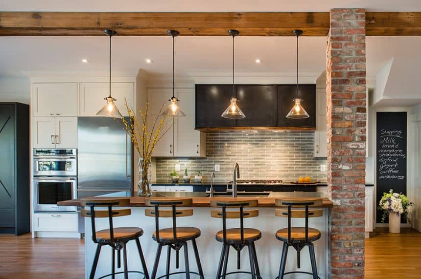 This industrial-style kitchen has a traditional-style twist to its white shaker cabinets and drawers of the kitchen island and peninsula. These are complemented by brick walls and pillars and an exposed wooden beam that supports pendant lights.