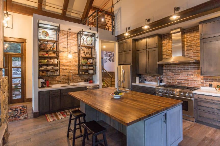 This industrial-style kitchen has a distinct charm to its red brick wall backsplash that complements the dark gray cabinets of the kitchen peninsulas that has matching dark gray floating cabinets and shelves as well as an exposed metal beam above the cooking area.