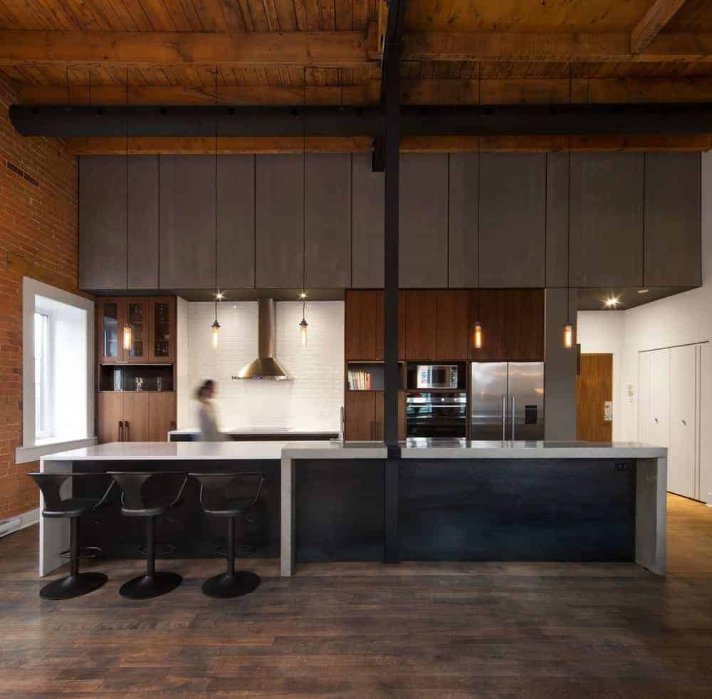 This view of the charming industrial-style kitchen shows a large kitchen island with waterfall white countertops and a black wooden facade pairing with the black stools. This also matches with the black support column and air duct of the high wooden ceiling that has exposed wooden beams.