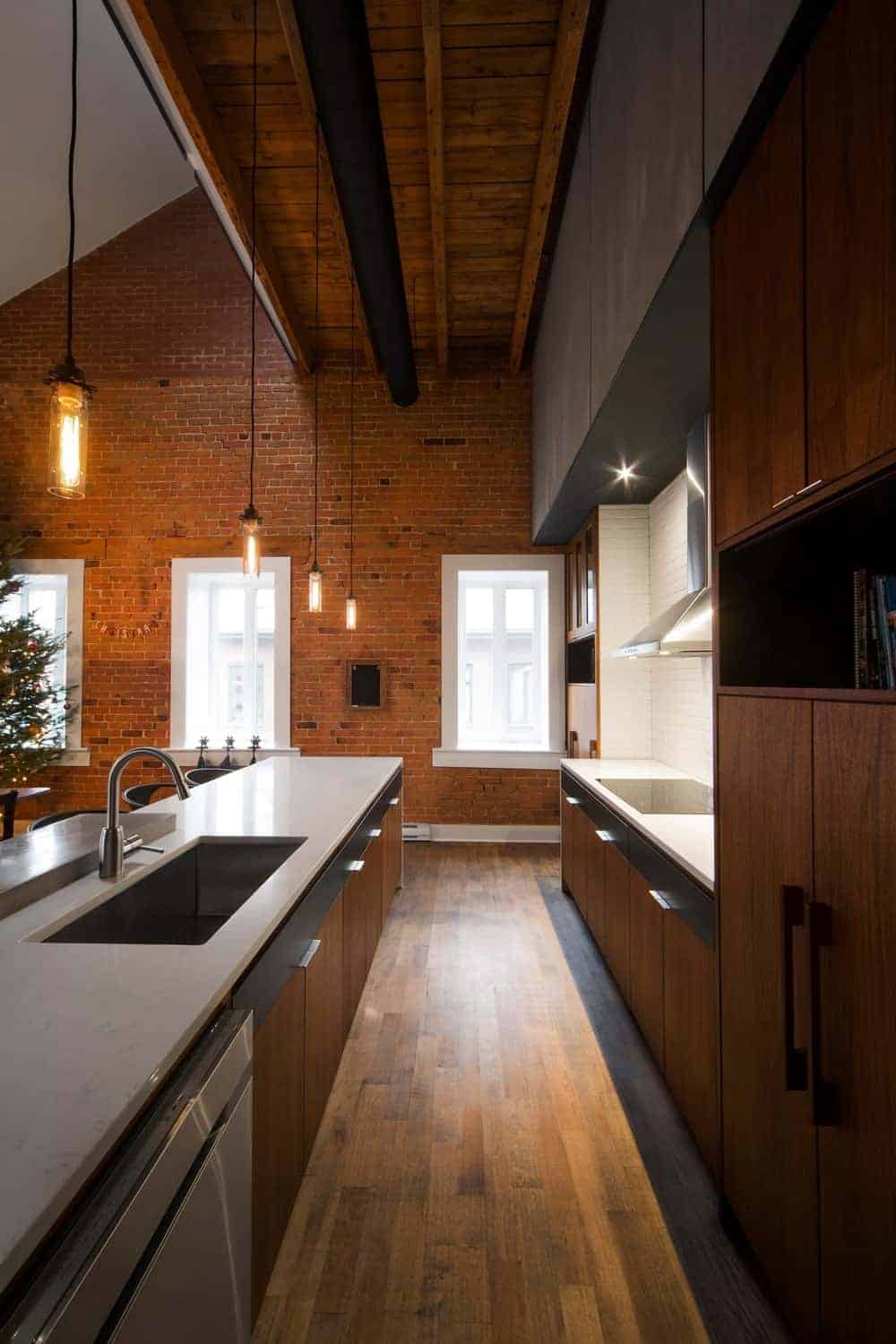 The narrow hardwood flooring of this Industrial-style kitchen matches with the brown wooden cabinetry of the flanking kitchen island and kitchen peninsula with white countertops and stainless steel appliances with a nice background of a brick wall with contrasting white windows.