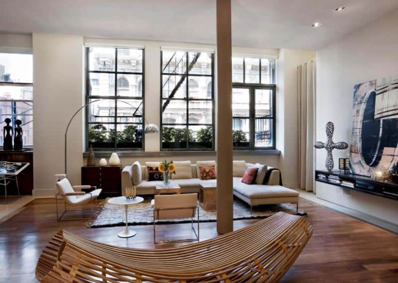 The high ceiling of this industrial-style living room is paired with a thin brown column that matches with the hardwood flooring that is brightened by the large glass windows behind the beige L-shaped sectional sofa paired with a modern standing lamp.
