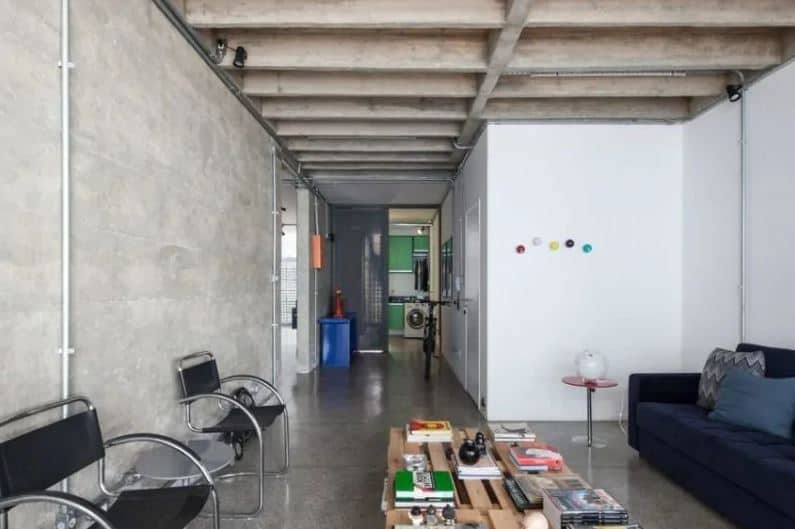 The dark velvet sofa is paired with a pallet crate coffee table and a couple of modern metal armchairs with black leather straps that stand out against the gray concrete walls adorned with exposed pipes and wiring.