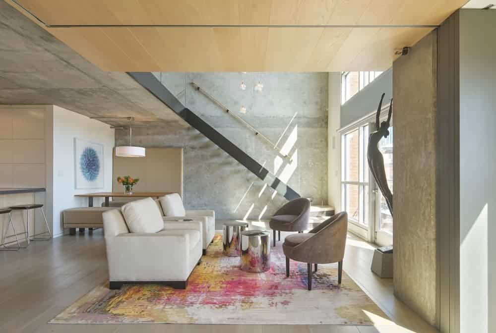 The colorful and chic painting-like large area rug that covers the hardwood flooring provides a nice dash of color for the industrial-style living room and its concrete ceiling and walls matching the cushioned chairs and white sofas.
