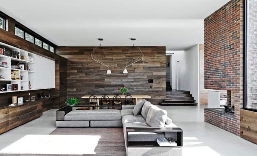 Behind the light gray L-shaped sectional sofa has a fireplace inlaid with a large column of red bricks. In front of the sofa is a large wooden structure that is embedded into the wall that houses cabinets and shelves filled with decors.