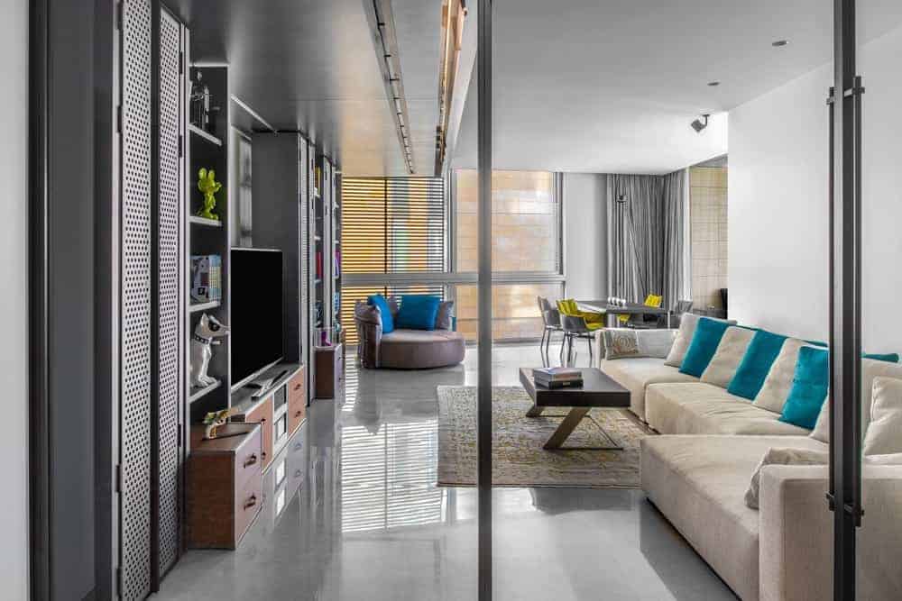A large velvet beige L-shaped sofa takes over most of the floor space of this industrial-style living room that has a large gray metal structure across from the sofa. This houses the entertainment system and has cabinets and shelves.