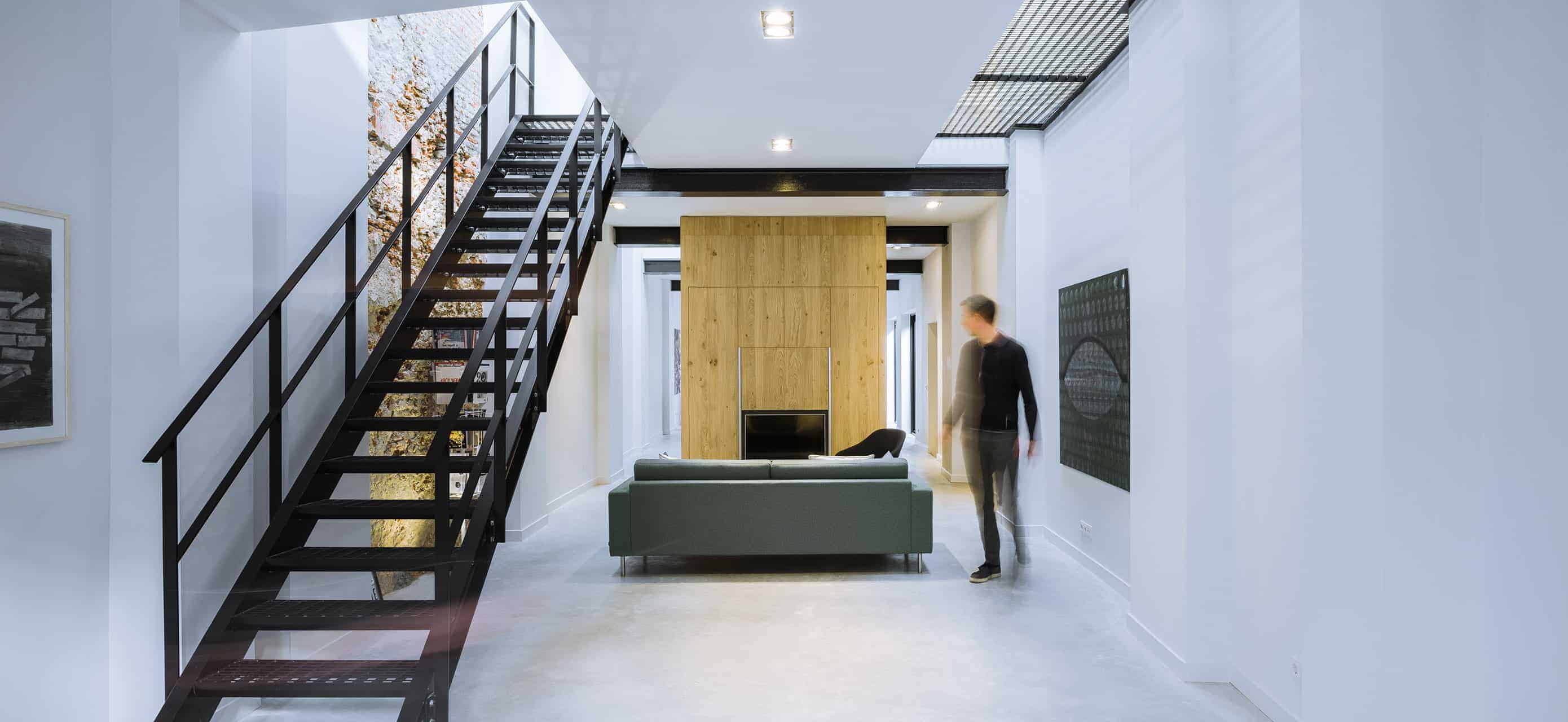 The white walls of this bright living room is a match for the white industrial-style flooring that contrasts the gray sofa facing a wooden wall panel that houses the fireplace. This brightness is contrasted by the dark metal elements of the stairs and the beams of the ceiling.