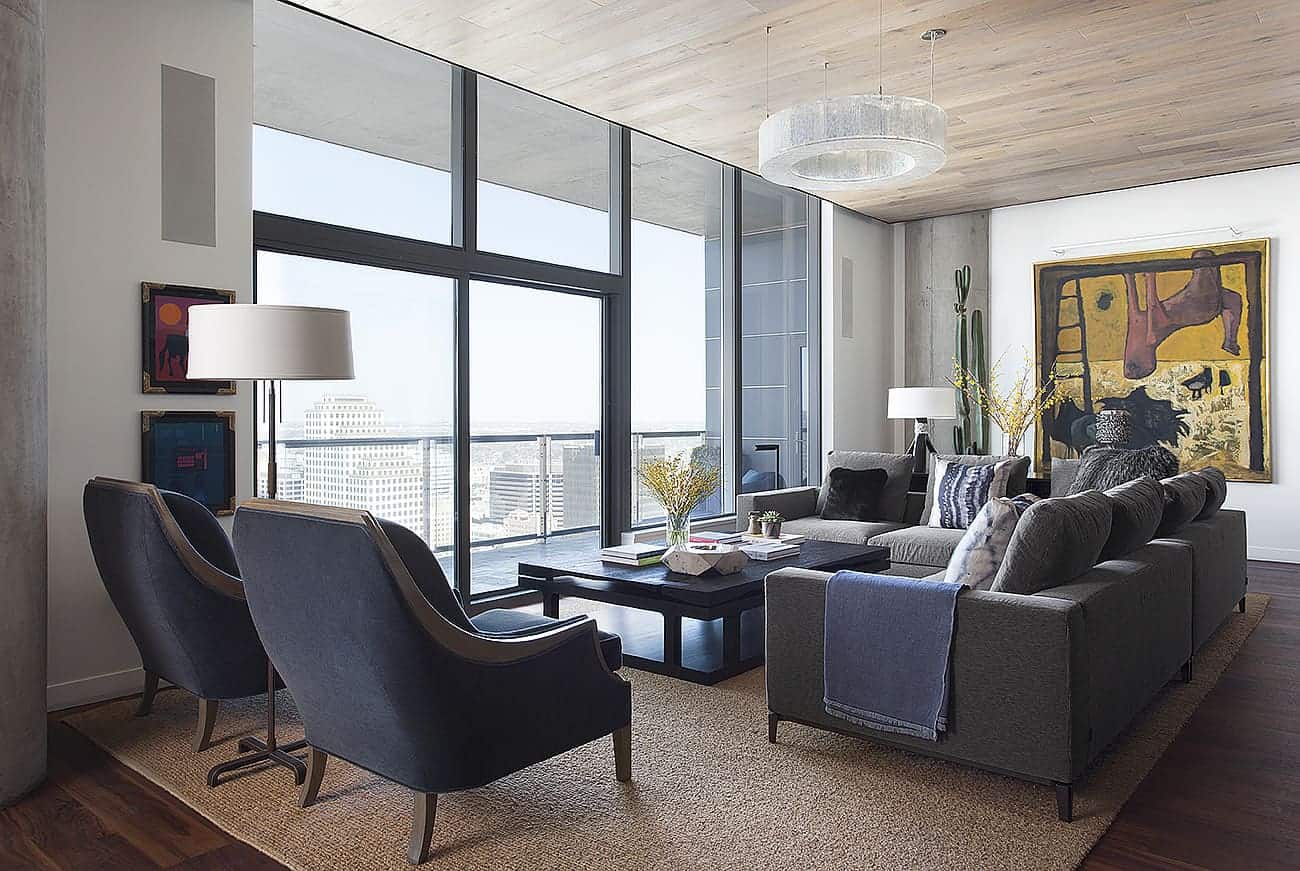 The white wooden ceiling that is brightened by the natural lights of the large glass wall is contrasted by the dark hardwood flooring that is topped with a large brown area rug paired with dark gray sofas and a couple of dark blue cushioned armchairs.