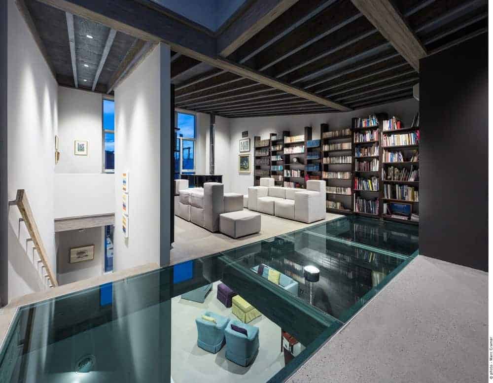 There is a unique glass flooring with a green tint that leads to this living room that is surrounded by multiple bookshelves that match the ceiling with a same dark hue of the shelves. This makes the modern white leather sofa stand out.