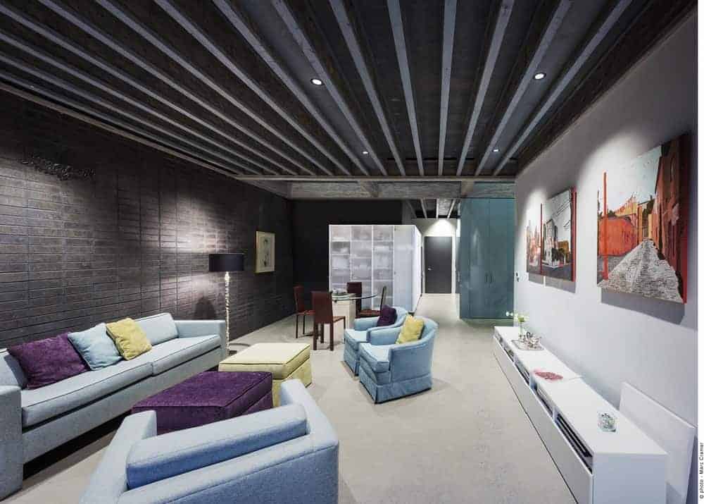 This living room has a wide ceiling filled with dark gray metal beams with recessed lights that complements the dark gray stone walls that contrast the light blue sofa set facing the concrete wall adorned with colorful paintings.