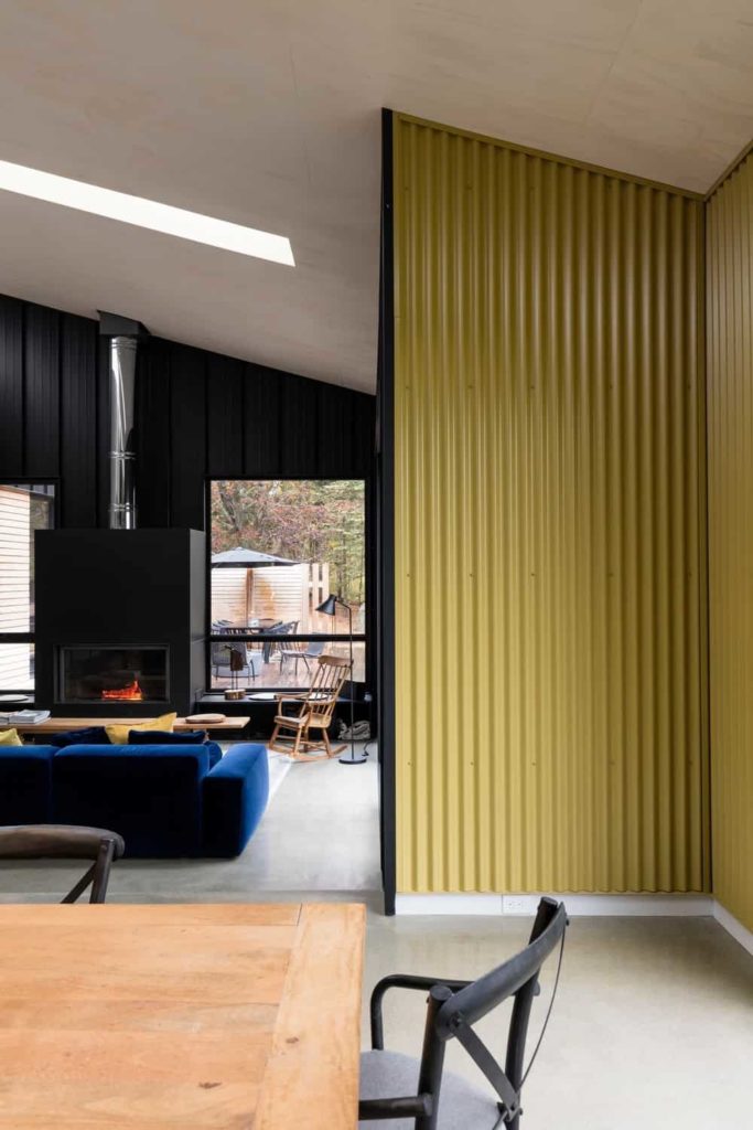 There are yellow patterned metal walls leading to this industrial-style living room that has black walls made of what looks like roofing materials. This matches with the black housing of the fireplace facing the blue velvet cushioned sofa.