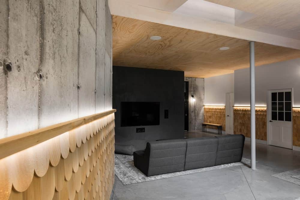 This simple living room has a dark gray sectional sofa facing a matching dark gray wall that supports the wall-mounted TV and its sound system. These gray elements stand out against the bare wooden ceiling that matches the bare wooden wainscoting with a textured scales design on it.