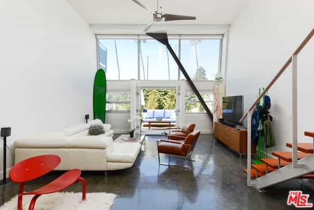 The dark gray industrial flooring of this living room contrasts the white L-shaped sectional sofa and the white walls that are brightened by the large glass wall of the entryway that has a large metal beam and double doors.