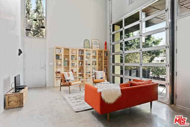 This living room has a bright orange cushioned sofa that stands out against the light hue of the industrial-style flooring and white walls that are adorned with a large bookshelf adjacent to a large roll-up glass door.