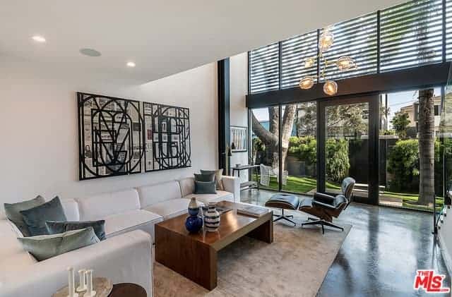 This simple white living room has an industrial-style background of the large glass wall of the entrance to this home. It has metal frames in a dark hue that stands out against the brightness of the natural lights and the nice landscaping outside.