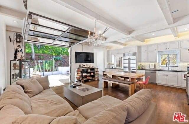 This simple and traditional-style living room by the kitchen is augmented with industrial-style elements. The white coffered ceiling that hangs a small white chandelier is accented with the roll-up glass door along with its wires and gears.