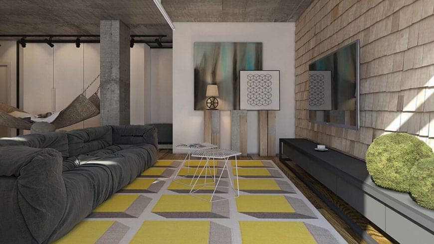 The yellow patterned area rug provides a bright colorful contrast to the gray sofa that matches with the gray concrete ceiling. This sofa faces a wall made of exterior wall shingles that provides a complex contrast to the mounted TV.