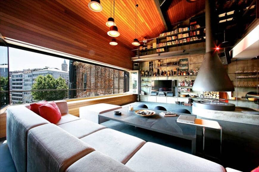 The wooden shed ceiling has a redwood shiplap finish to it that extends to the wall that is dominated with a wide and narrow open space thanks to folding glass windows. This brings in natural lights to the light cushioned sofa and the dark flooring.
