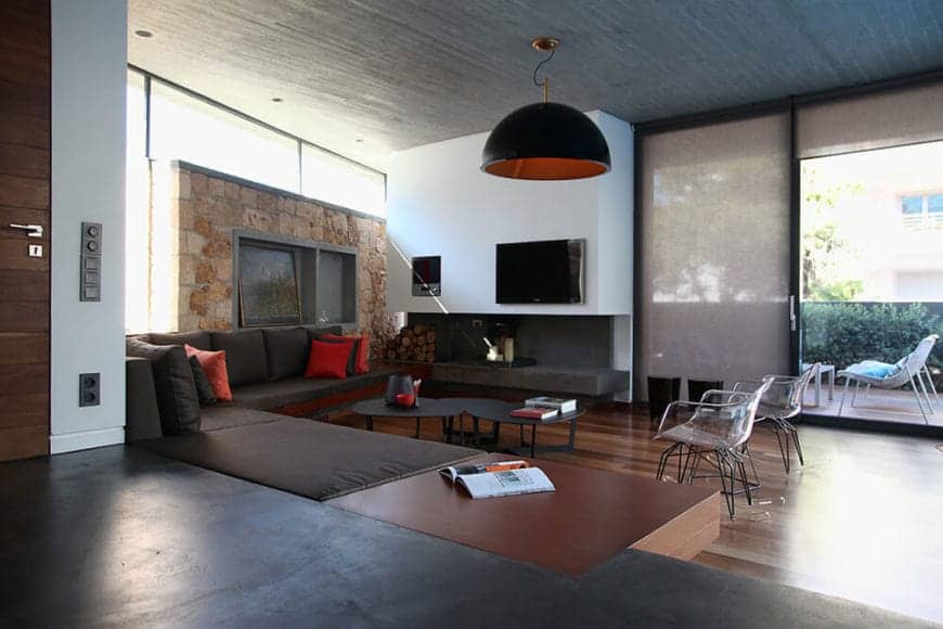 The large L-shaped dark brown cushioned sofa follows the lay of the walls with one wall made of beige stone while the other is a smooth white that houses the TV and the fireplace. These are all under a high wooden shed ceiling.