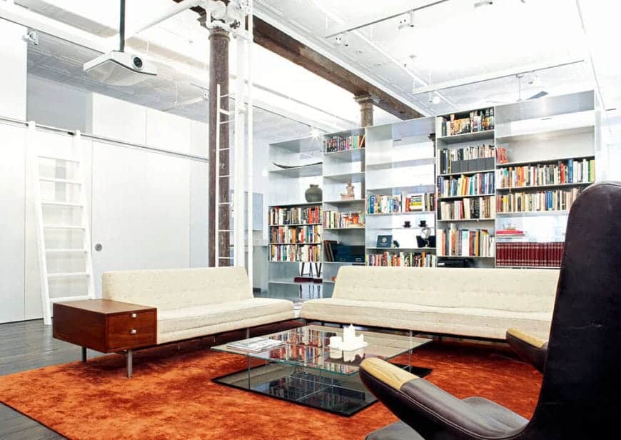 The bright orange velvet area rug stands out against the hardwood flooring and the white walls and ceiling. This ceiling has exposed pipes and wiring as well as a projector above the beige cushioned sofas facing a glass-stop coffee table.