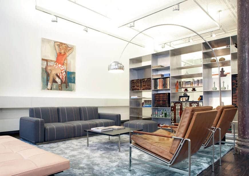 The high white ceiling is filled with exposed pipes, tubing and wiring for the brilliant white lights that make the ceiling and walls glow. These walls are adorned with a large painting and a large open shelving that gives the cushioned sofa set a nice complex background.