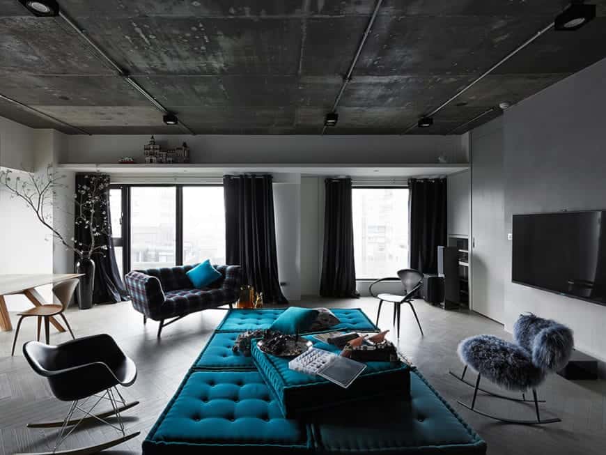The gray concrete ceiling has exposed wiring for its flush-mount lights over the green tufted day bed of the living room with a gray concrete flooring and light gray walls adorned with the dark curtains of the large glass windows and doors.