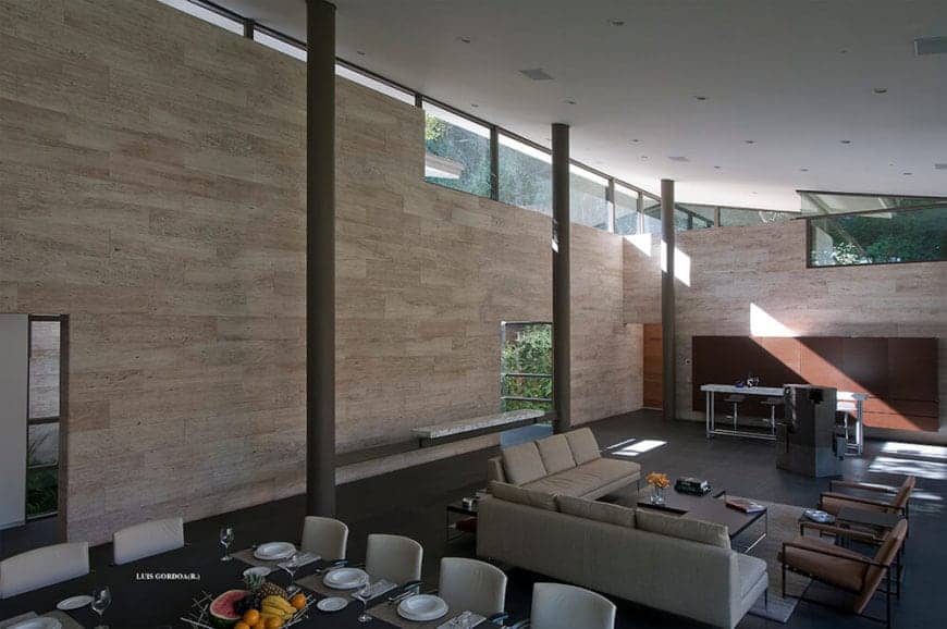 This large hall with a high white shed ceiling houses the dining area and living room area on the same dark gray flooring that matches the tall columns and contrasts the light gray sofa set and the light gray area rug.