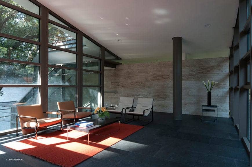 The stand-out elements of this living room are the orange area rug over the industrial-style stone flooring and the matching orange cushioned armchairs against the gray metal frames of the glass wall extending to the white shed ceiling.