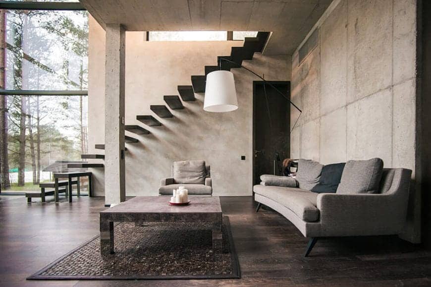 This living room is in a large alcove made of gray concrete ceiling and walls paired with a dark hardwood flooring that is contrasted by the light gray curved sofa and a cushioned armchair facing a rectangular coffee table.