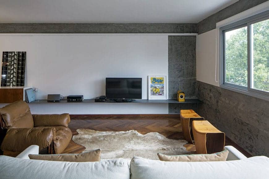 This simple living room has a brown leather cushioned armchair that matches with the hardwood flooring with a herring bone pattern. This is contrasted by the white walls accented with sections of gray concrete.