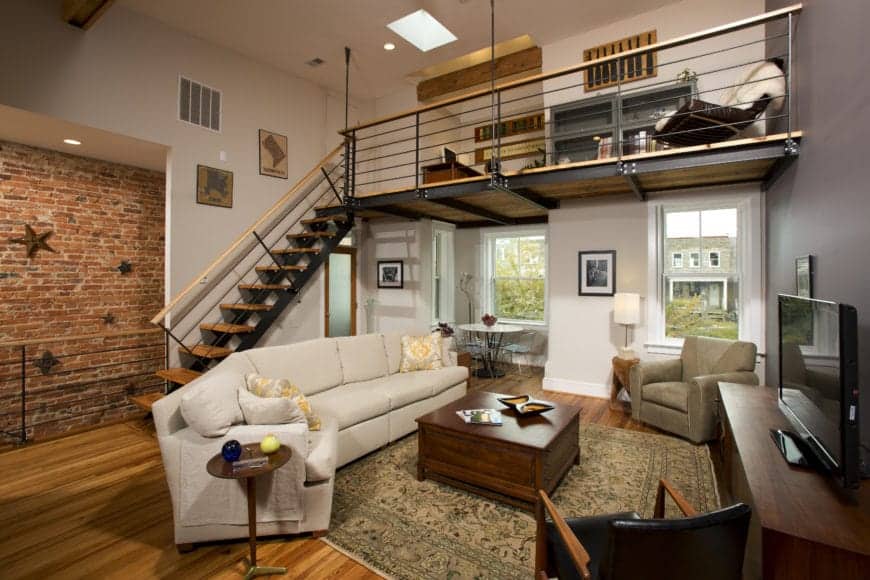 This appears to be a traditional-style living room that is framed with industrial-style elements like the red brick wall adorned stars, the metal staircase leading to a metal loft above that forms a small ceiling above the living room with exposed metal beams.