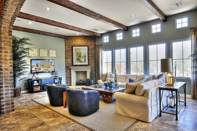 The bright row of windows on one side of this living room brings in an abundance of natural lighting to the white ceiling and its exposed wooden beams as well as the adobe bricks of the fireplace and the arched entryway.