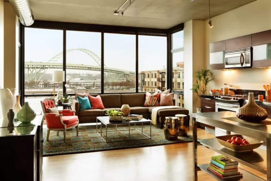 The L-shaped sectional sofa has colorful pillows that match the tufted cushioned armchair beside it. This is given a nice urban background featured by the large glass wall that illuminates the concrete gray ceiling and its exposed vent.