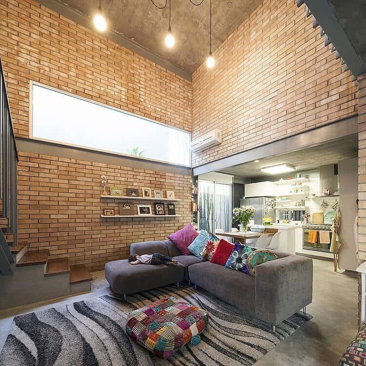 The gray L-shaped sectional sofa stands out against the red brick walls extending all the way up to a high concrete ceiling adorned with a brilliant transom window that augments the warm yellow lights of the pendant lights.