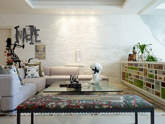 The white tray ceiling of this living room is contrasted by the texture of the white stone wall with a brick wall pattern to it. This is adorned with artworks above the beige L-shaped sectional sofa facing a large glass-top coffee table.